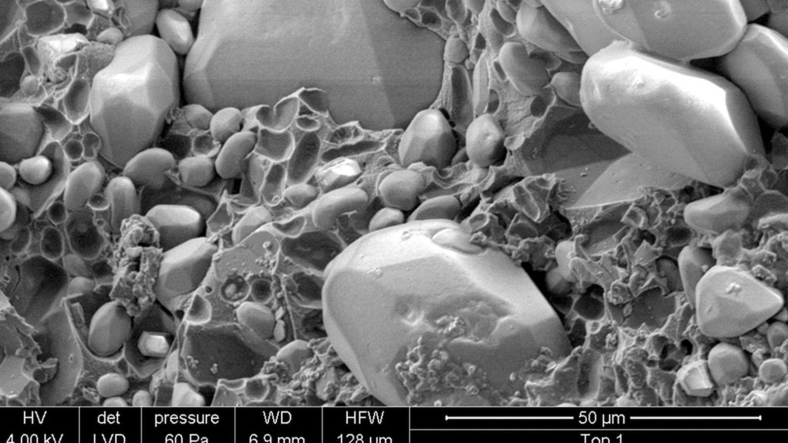 Microscopic image shows a highly magnified view of mineral particles, including smooth round grains and irregularly shaped fragments, with scale indicators and technical details at the bottom.