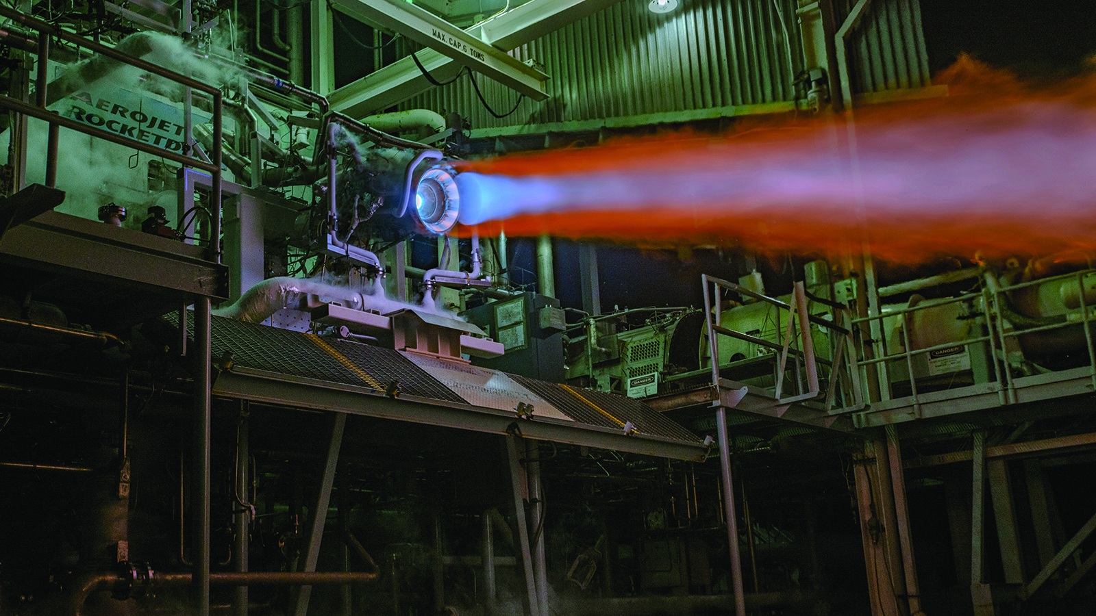 A rocket engine emits a bright flame during a test at a facility filled with industrial equipment and structures.