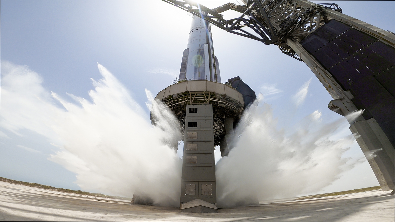Rocket on launch pad with water jets spraying at the base during a test, under a clear blue sky.