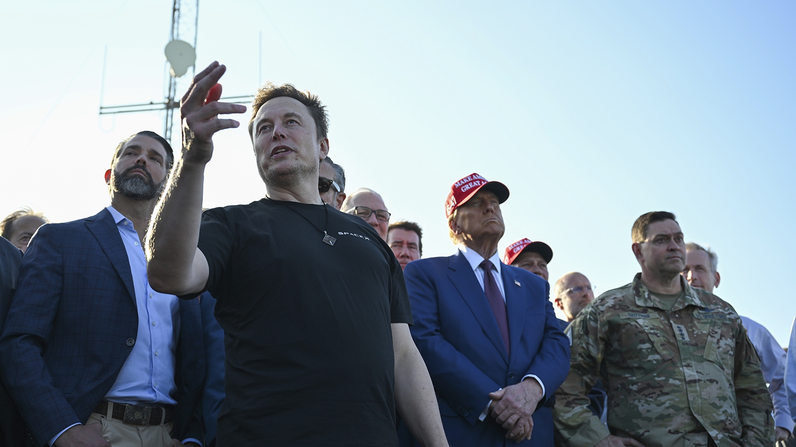A group of men stand together outdoors. One man in a black shirt gestures with his hand, while another in a suit and red cap looks on. Military personnel are also present.