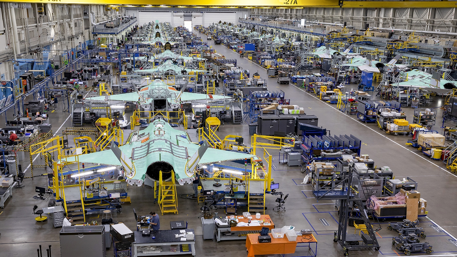 A large factory floor with multiple aircraft under construction, surrounded by various workstations, machinery, and tools. The aircraft appear to be in various stages of assembly.