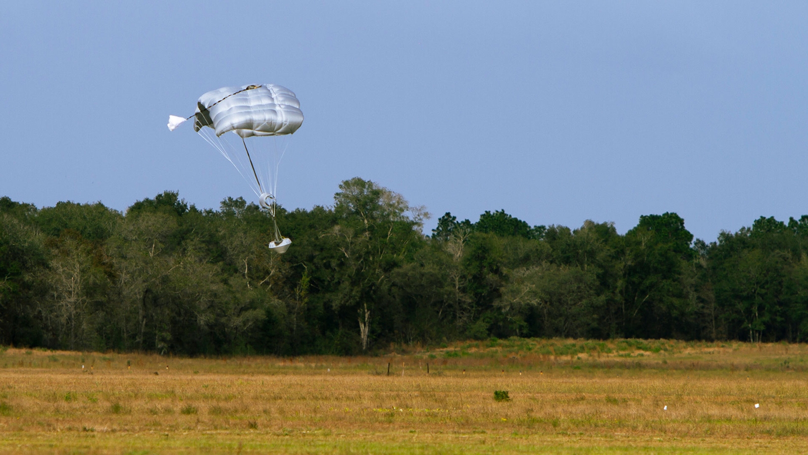 A white parachute descends toward a grassy field, with trees in the background and a clear blue sky overhead.