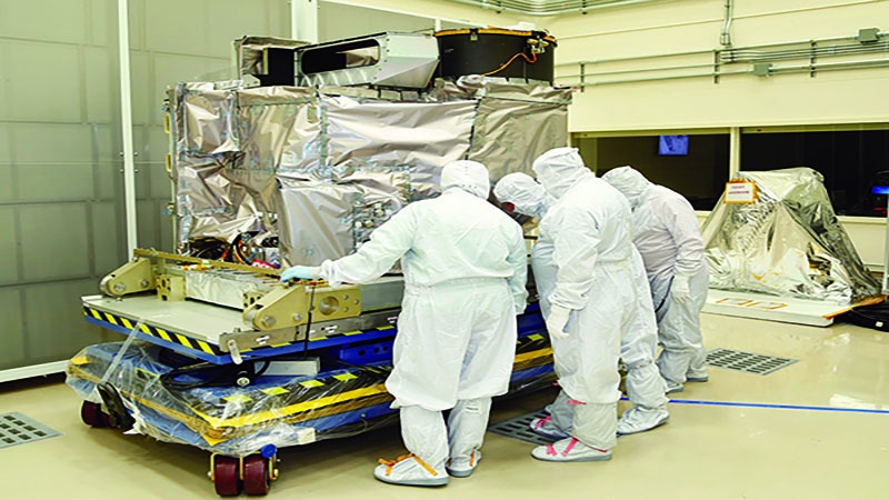 Three technicians in cleanroom suits inspecting a large, covered spacecraft component in a laboratory.