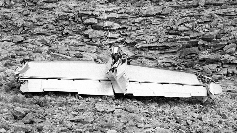 Black and white image of a crashed airplane wing lying on rocky terrain, with a person inspecting the wreckage.