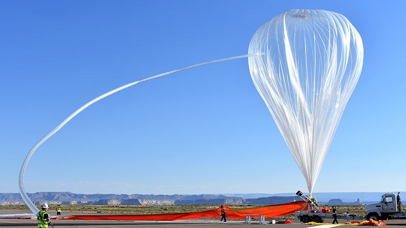 A large, translucent high-altitude balloon is being launched from a desert base, with ground crew members managing the equipment.
