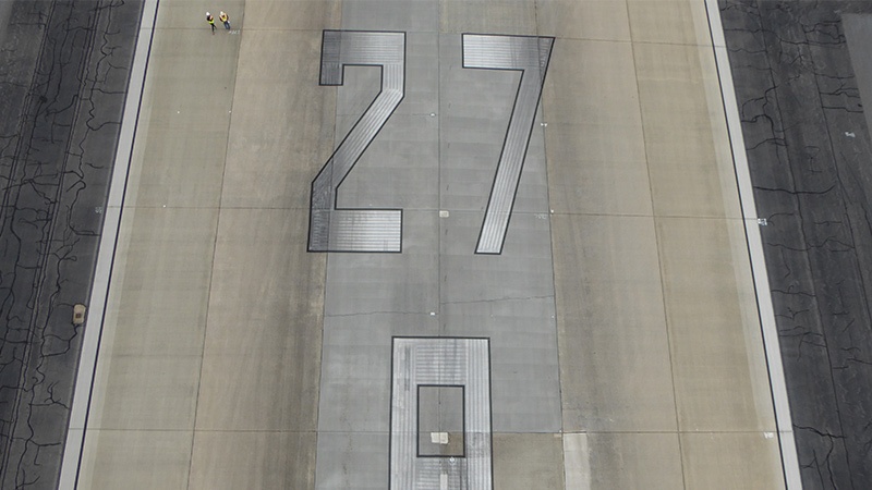 Aerial view of the number 27 painted on a cracked airport runway surface with a person standing next to it.