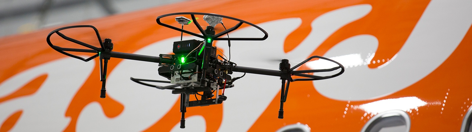 A small drone with green lights flies in front of an orange backdrop with white circular patterns.