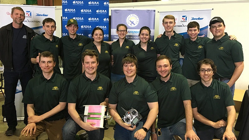 A group of people, mostly in matching green shirts, pose for a photo in front of banners. Some are holding objects while others stand or kneel in the background.