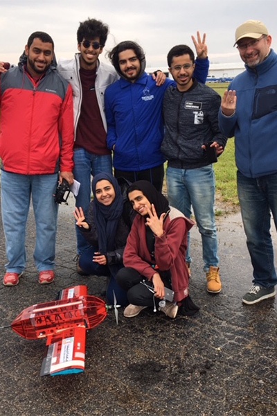 A group of seven people, some standing and some crouching, pose on a paved path outdoors. They smile and gesture while behind a small model aircraft on the ground. It appears to be a cloudy day.