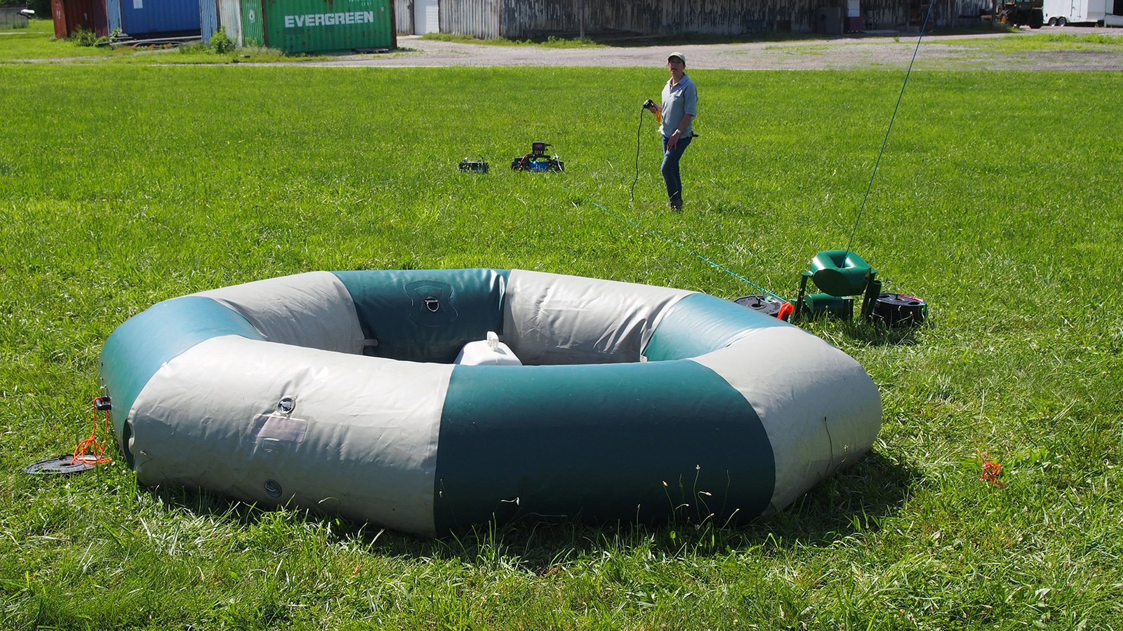 A person stands on a grassy field near an inflatable, gray and green circular structure, with equipment and bins nearby. Containers and a small vehicle are in the background.