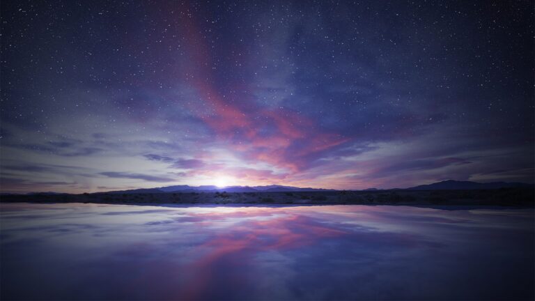 A serene night sky filled with stars over a calm body of water reflecting the faint glow and colorful hues of the sunset or sunrise on the horizon.