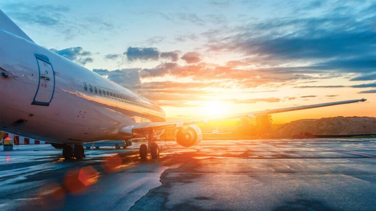 May 2019 AIAA Bulletin A commercial airplane is parked on a wet tarmac at sunrise, with the sun shining through scattered clouds in the background.