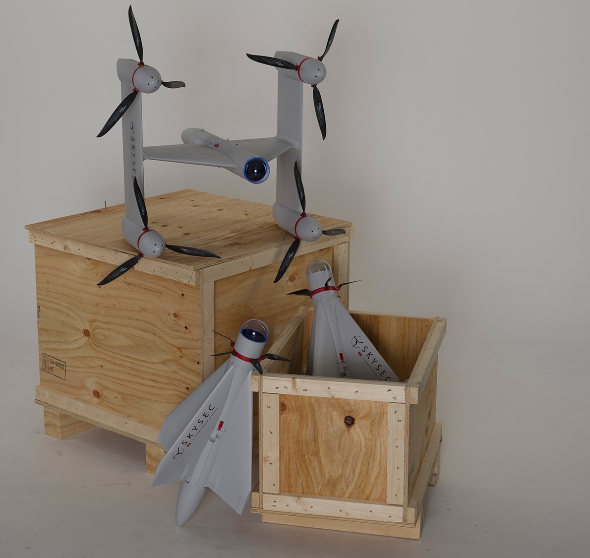 Two wooden crates on a white background, with a drone placed on top and two small rocket-like objects partially inside the smaller crate. Both crates are open.