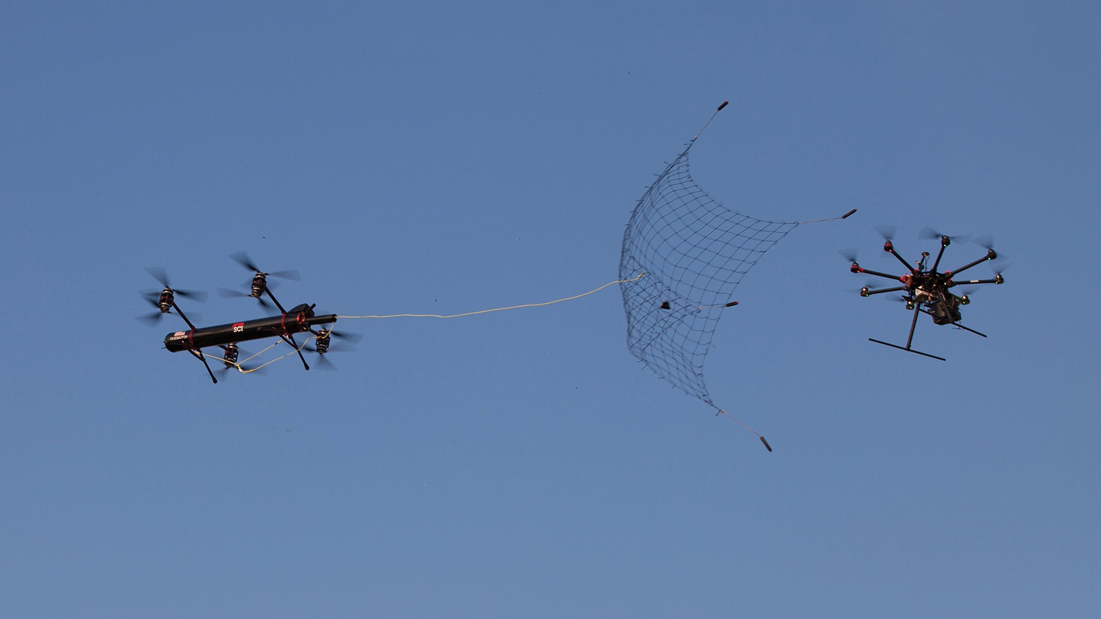 Two drones in the sky; one drone is pursuing the other while deploying a net to capture it. The background is clear and blue.
