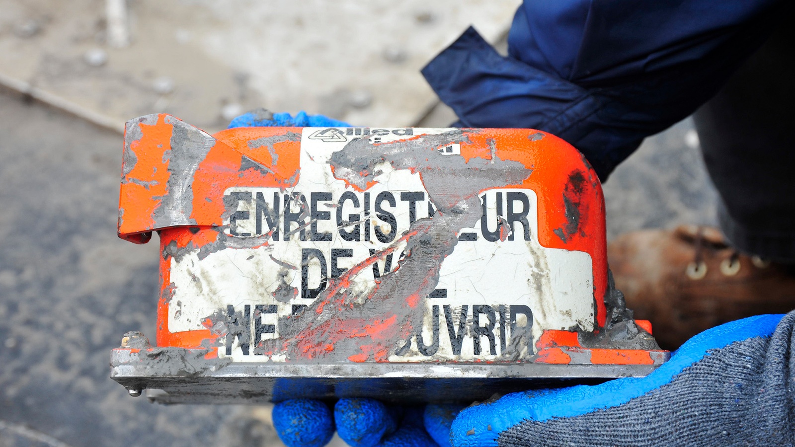 Close-up of a person holding a flight data recorder, partially covered in dirt with text that reads