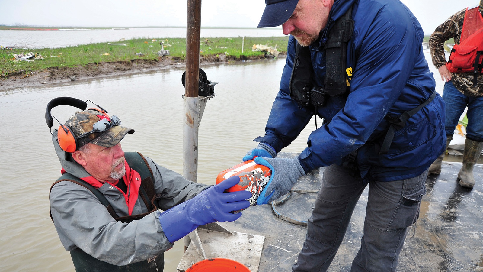 Two men work together to recover a cylindrical object from water, wearing protective clothing and gear. One is kneeling, the other standing, on a wet surface with water and grassy land in the background.