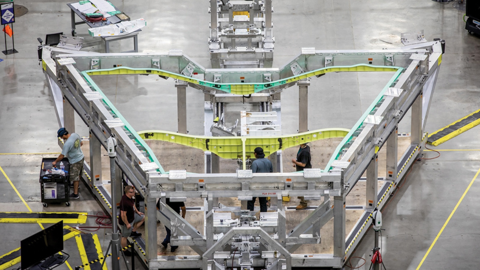 Workers assemble a large metallic framework in a factory setting. The structure has a V-shaped design with green inner sections, surrounded by various tools and equipment.