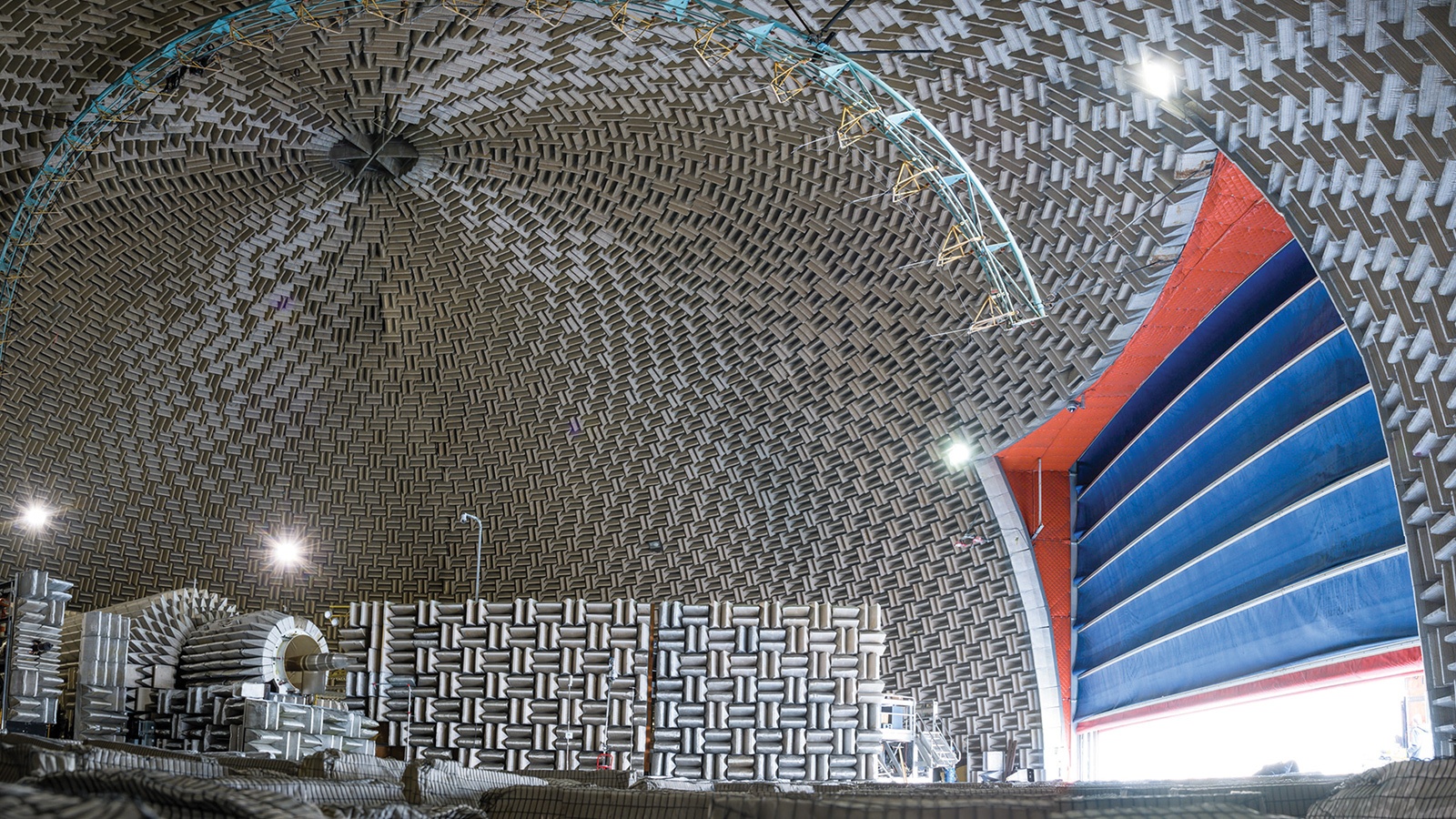 A view of an anechoic chamber with sound-absorbing panels covering the walls and ceiling. Large blue doors are partially open, revealing the inside of the chamber. Various equipment is visible on the left.