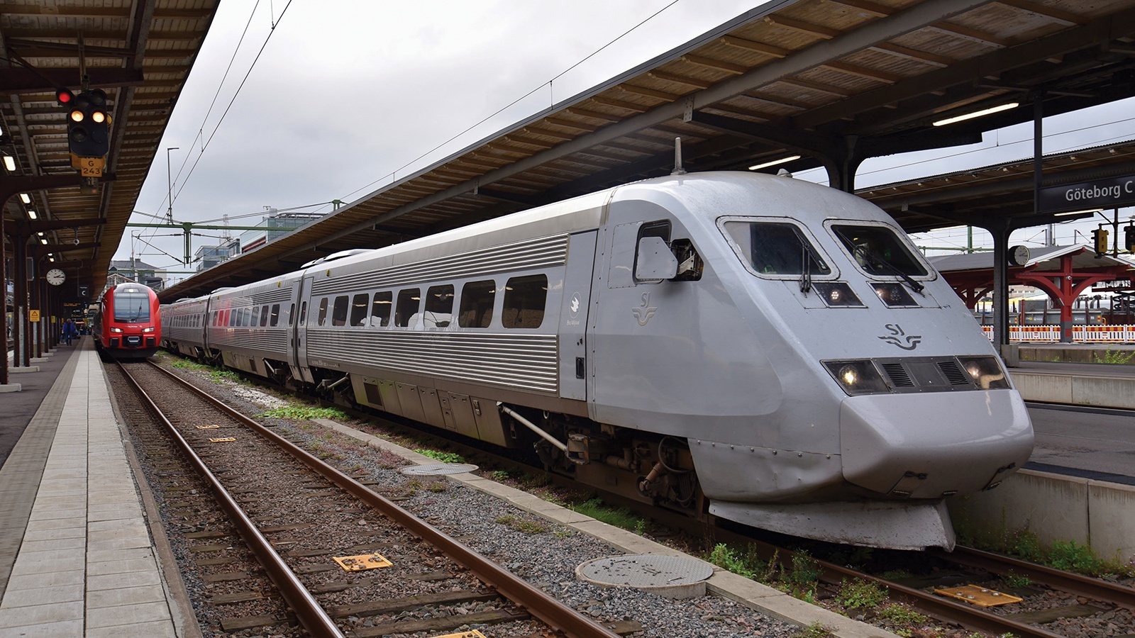 A silver train is stationed at a platform under an overhang, with another red and white train visible on an adjacent track. The platform has signs and a clock.