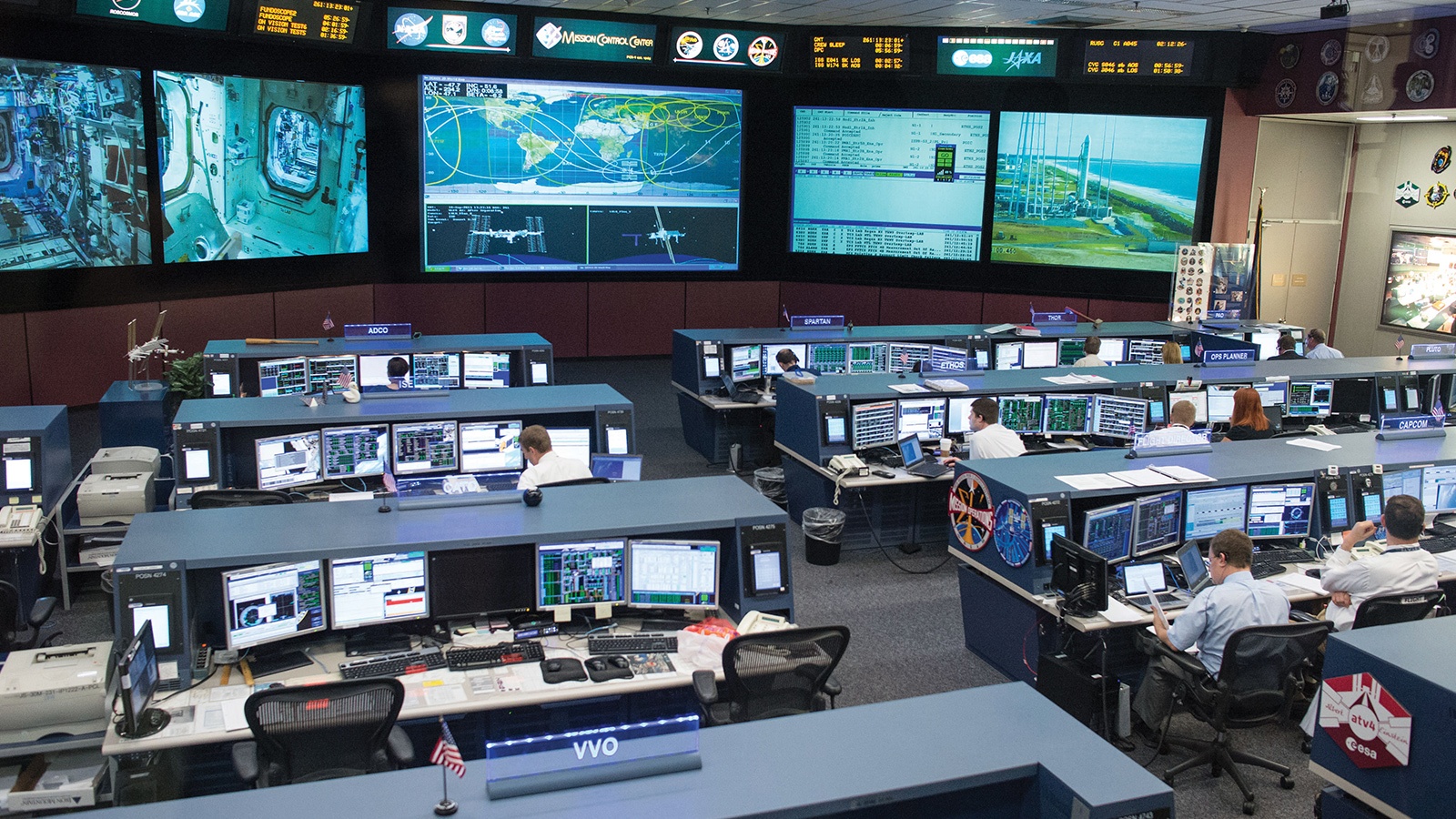 A spacious control room with multiple workstations and large screens displaying spacecraft data and missions. Several people are seated at their desks, monitoring and operating equipment.