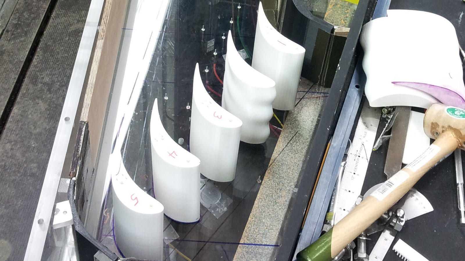 A row of white curved objects, each labeled with a red letter, are placed on a reflective surface near a cluttered workbench with tools and papers.