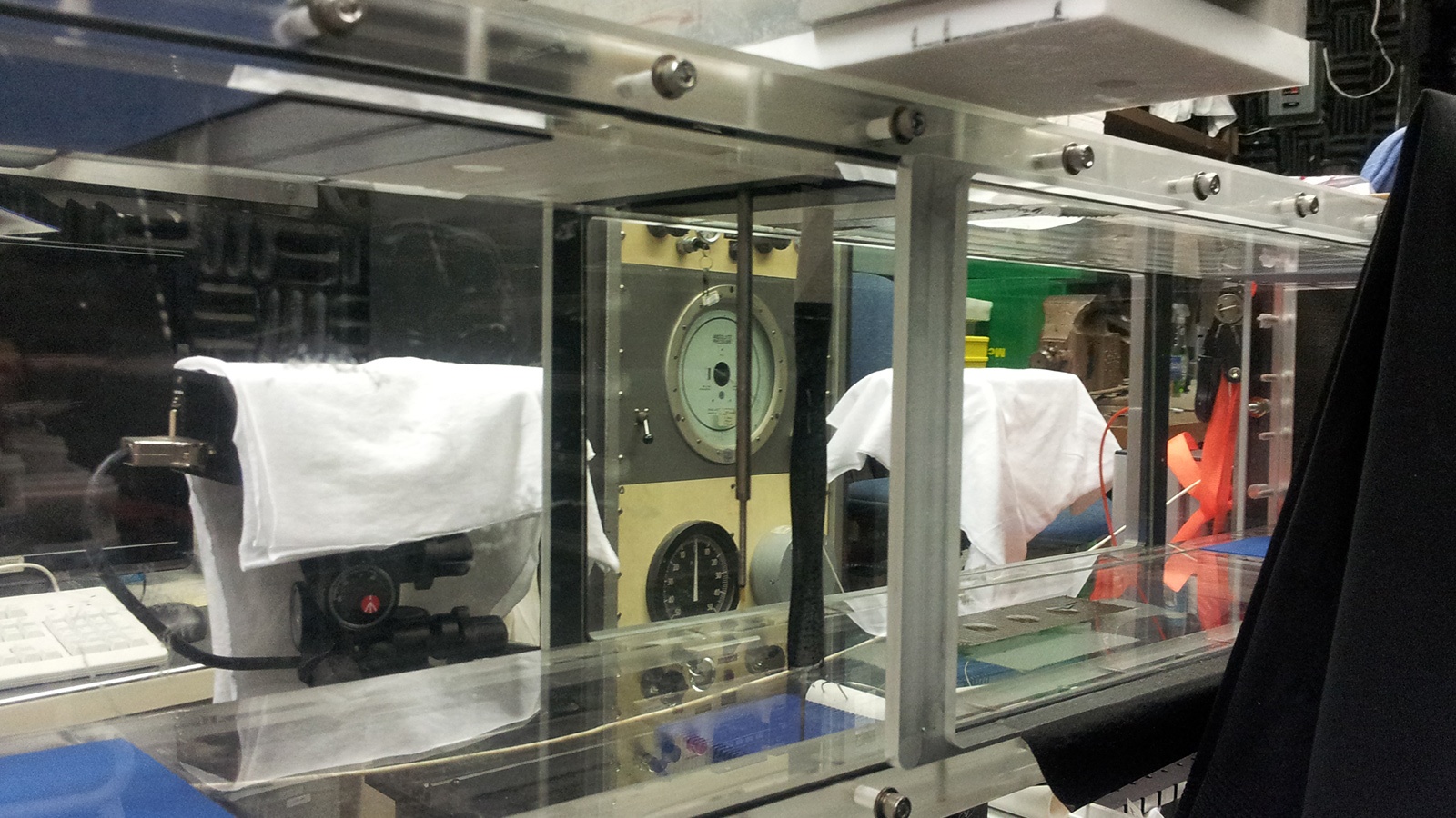 A transparent plastic enclosure with various instruments and gauges inside, including a large round dial. Textiles are draped over some of the instruments.
