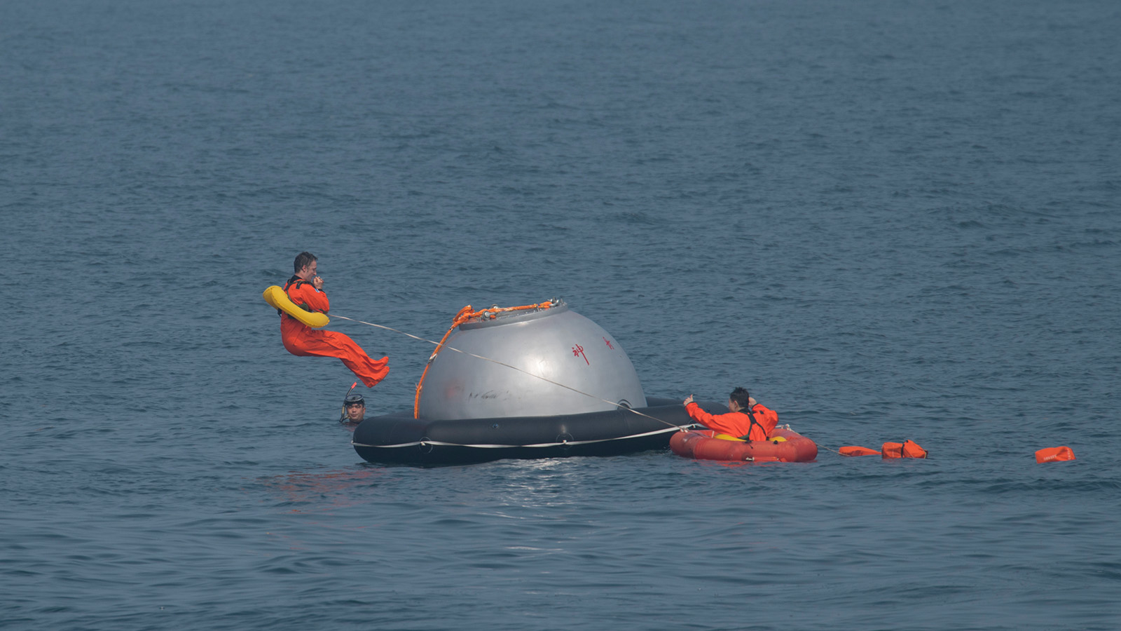 Three individuals in orange suits are in the ocean with a silver capsule. One person climbs onto the capsule, another holds a life ring, and the third is in an inflatable raft.
