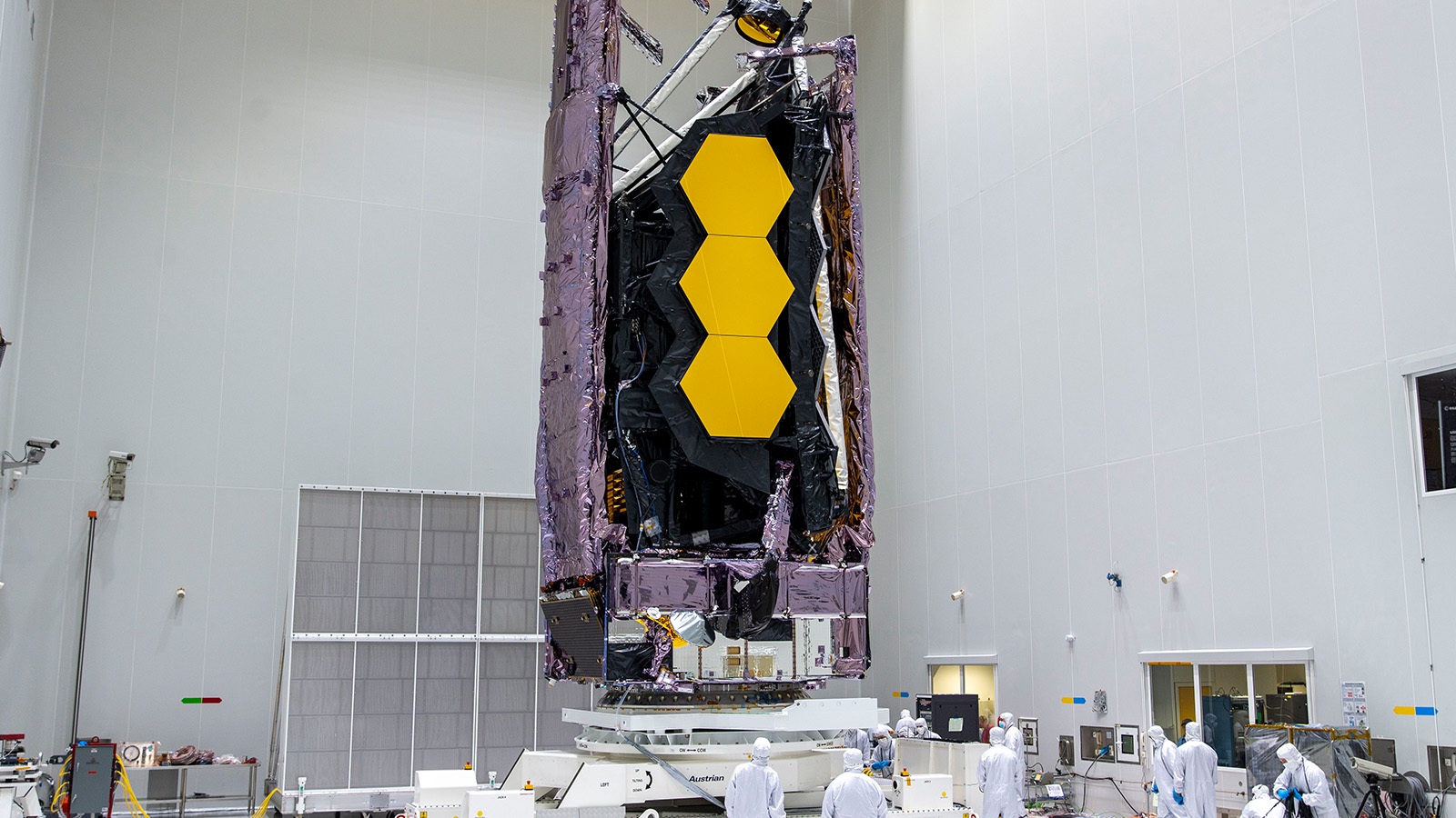 Engineers in white lab coats work around a large, gold-and-black space telescope, which is suspended vertically in a cleanroom.
