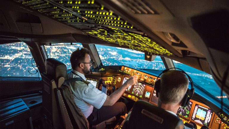 February 2022 AIAA Bulletin Two pilots, one in uniform, navigate a commercial airplane over a city at night, illuminated cockpit controls and exterior lights visible through the windows.