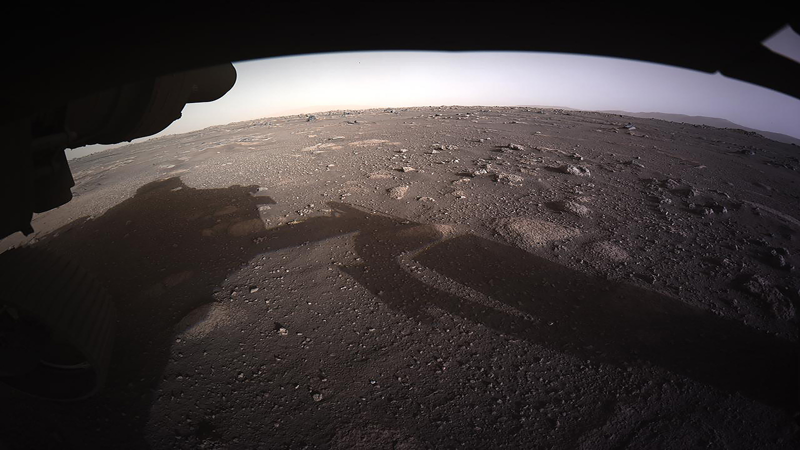 A robotic rover's shadow is cast on a rocky, dusty, and barren Martian landscape under a pale sky.