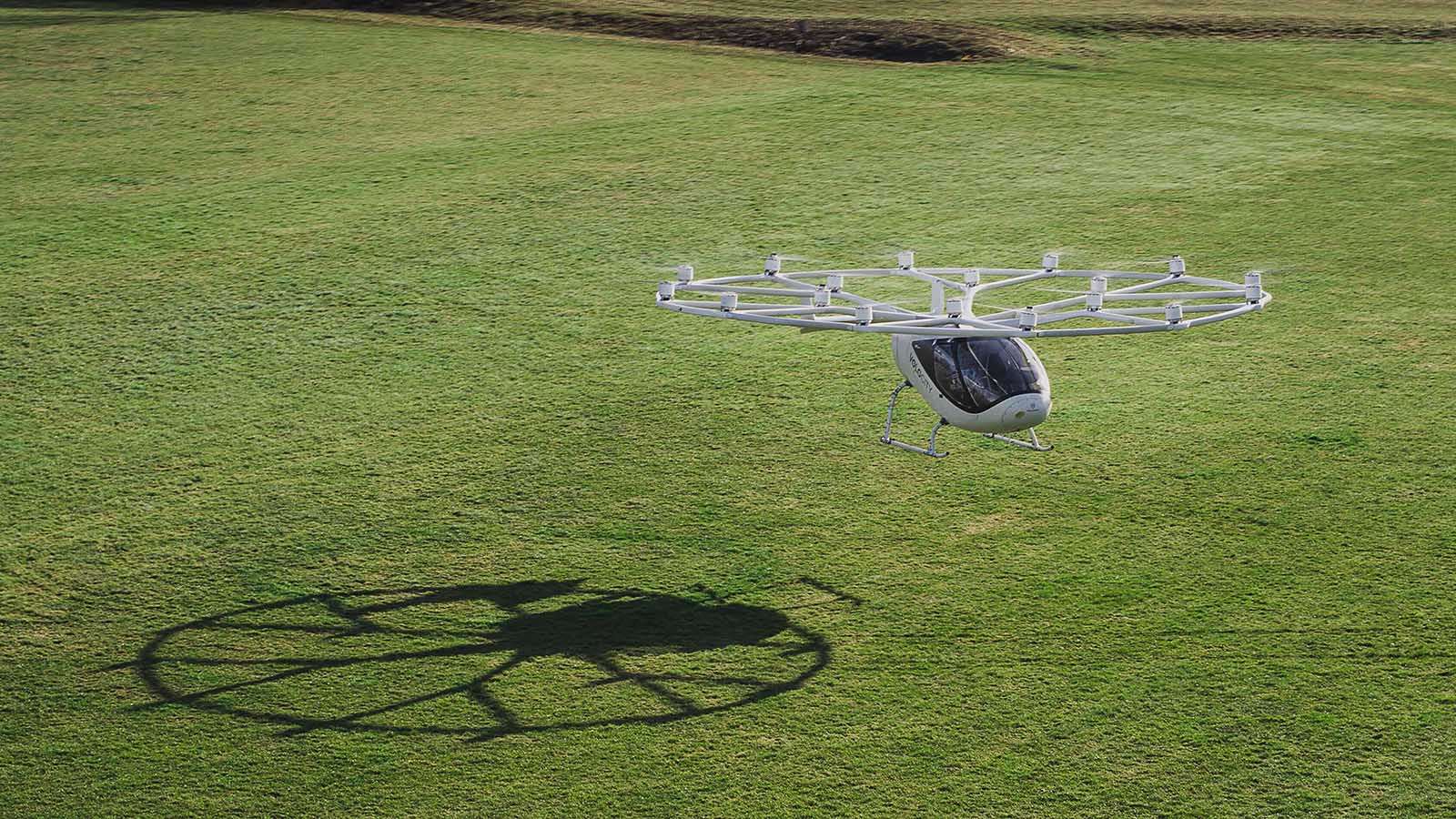 A white, multi-rotor drone-like aircraft hovers above a grassy field, casting a circular shadow on the ground below.