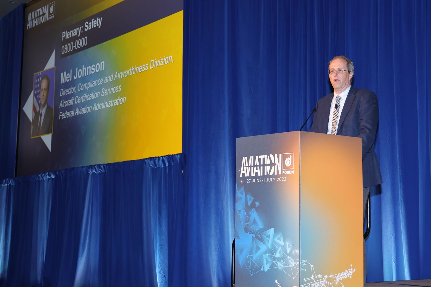 A man stands at a podium speaking at an aviation forum. A large screen displaying information about the event and the speaker,