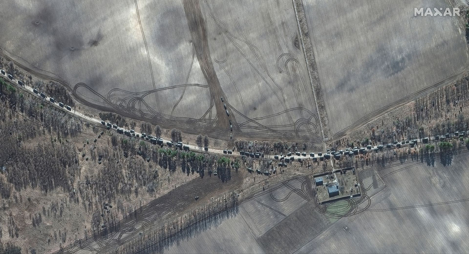 Satellite image showing a long line of military vehicles on a dirt road, surrounded by expansive fields with tire tracks and patches of trees. A small complex of buildings is visible on the bottom right.