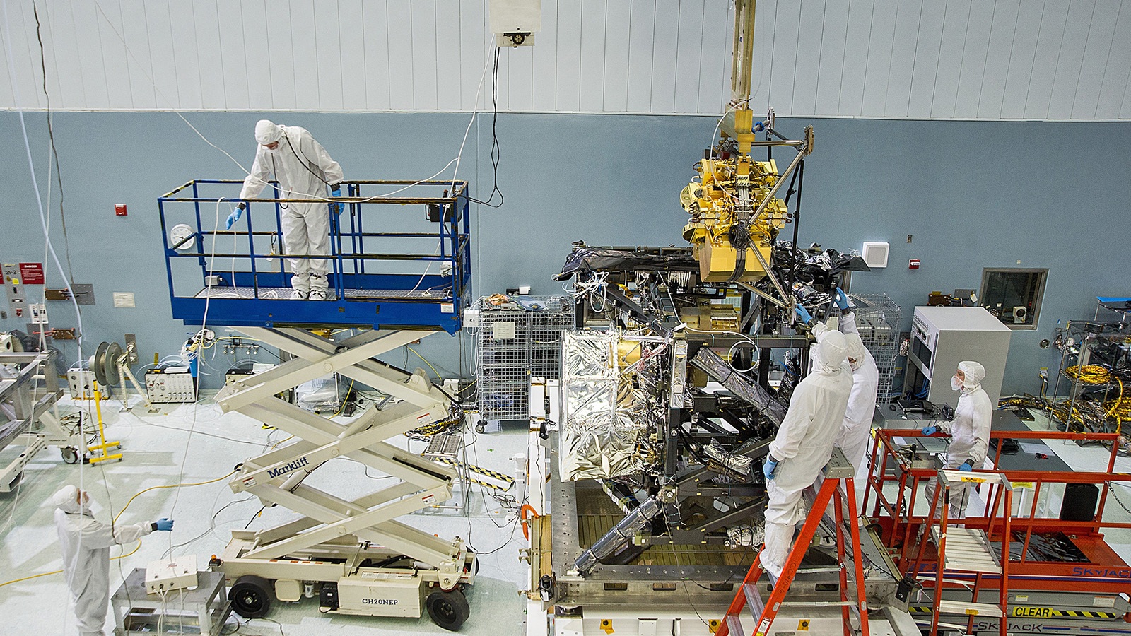 Technicians in cleanroom suits work with equipment and machinery in a laboratory. One person stands on a scissor lift, while others handle a large mechanical structure.