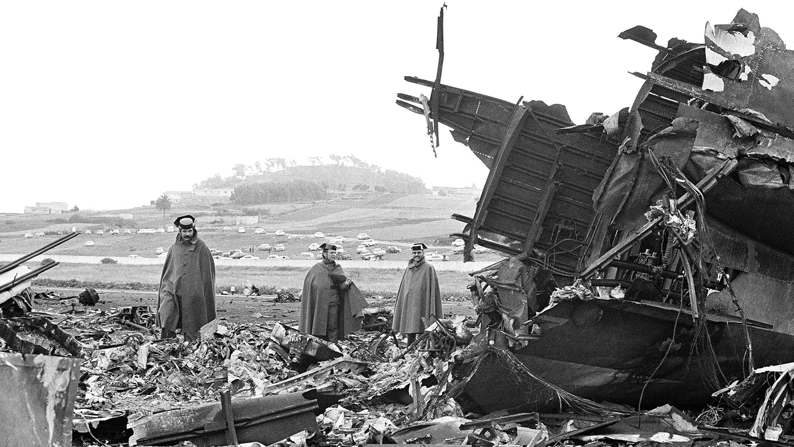 Three individuals in cloaks stand among debris and wreckage in a field, with a hill and several vehicles visible in the background.