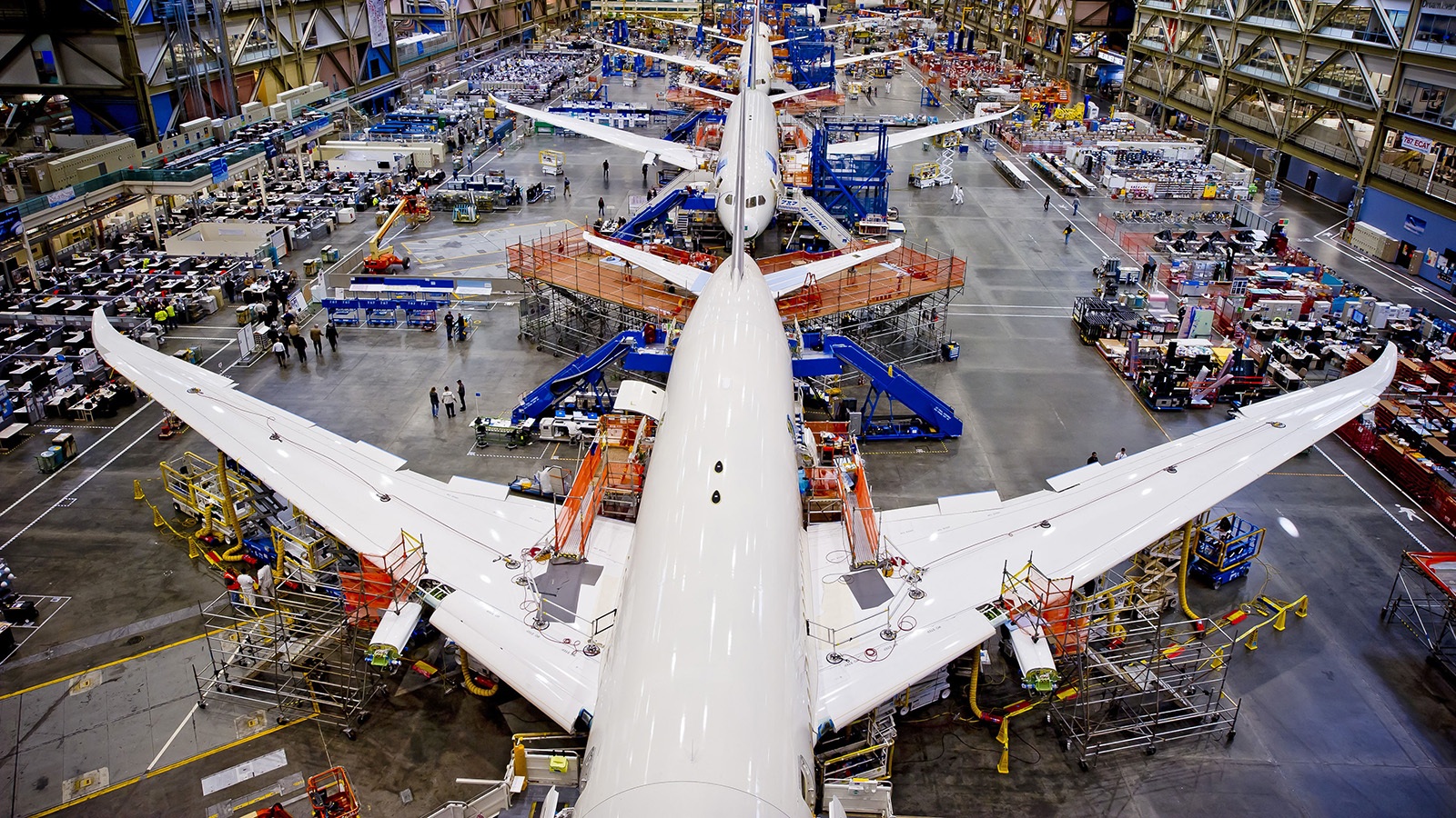 Wide-angle view of a large airplane manufacturing facility showing multiple aircraft under construction, surrounded by scaffolding, machinery, and workers throughout the spacious industrial environment.