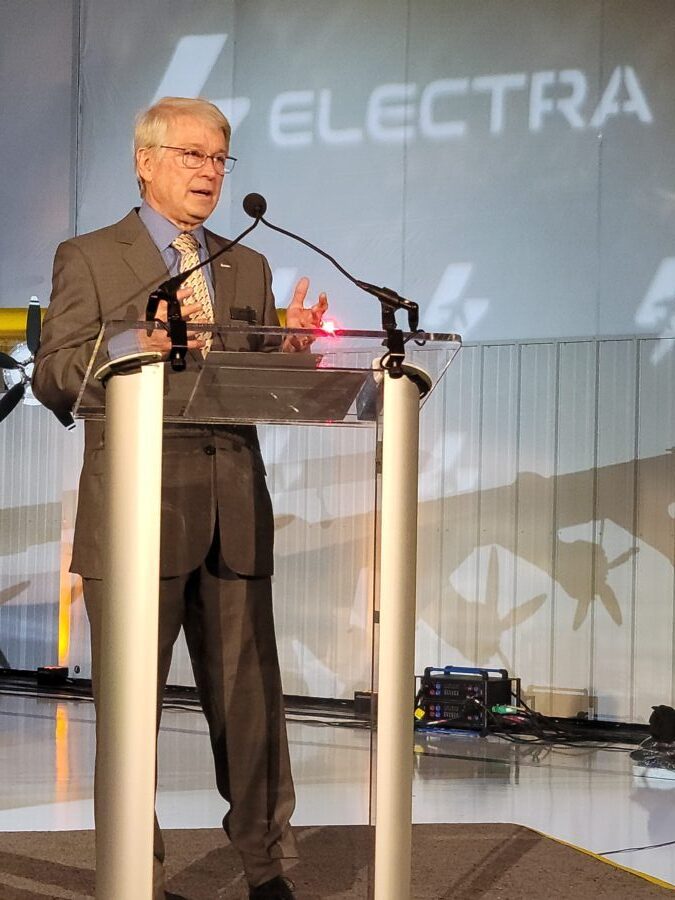An older man in a suit and tie speaks at a clear podium with microphones. The background features the word