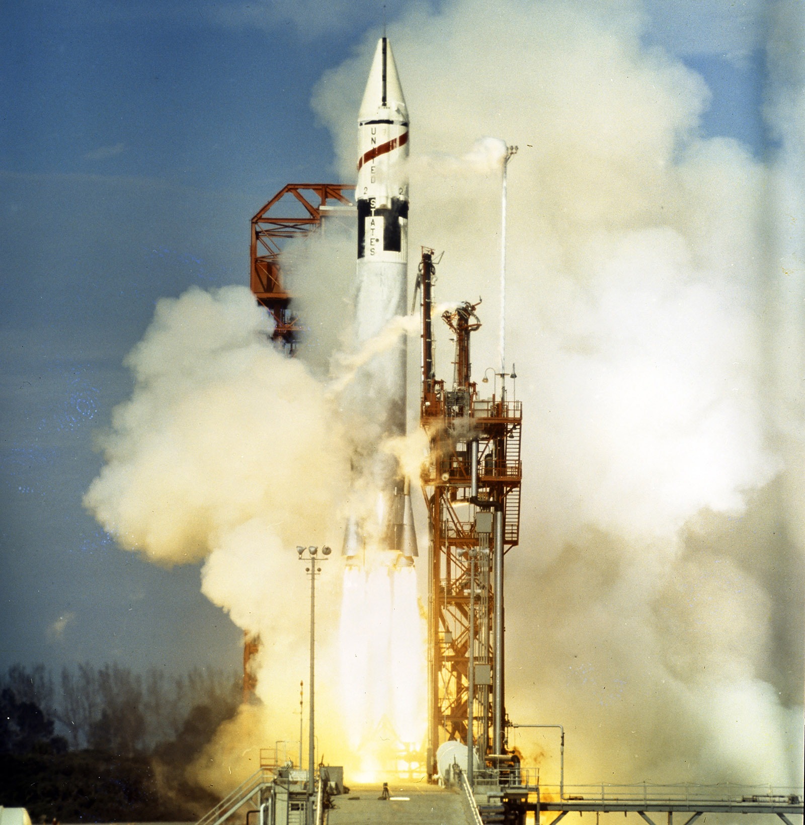 A rocket is launching from a platform, surrounded by smoke and flames indicating lift-off. The sky is clear and blue in the background.