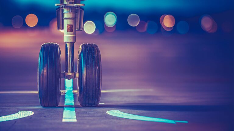 Close-up of an airplane's landing gear on a runway at dusk, with bokeh lights in the background.