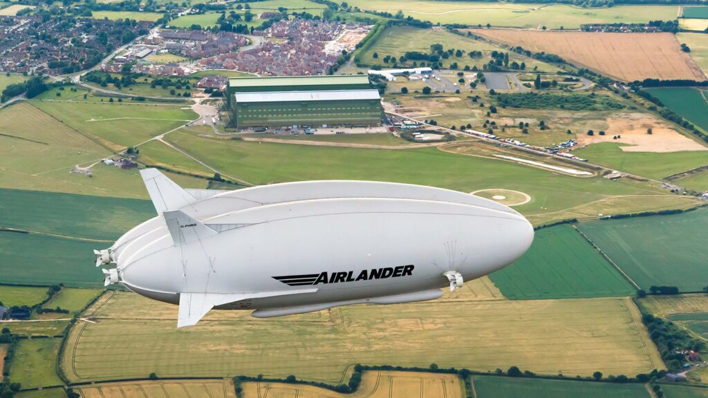 An Airlander blimp with "AIRLANDER" logo is flying above a rural landscape with fields, roads, and buildings visible below.