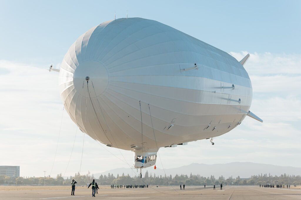 A large white airship is tethered on an airfield, with several people standing nearby under a clear blue sky.