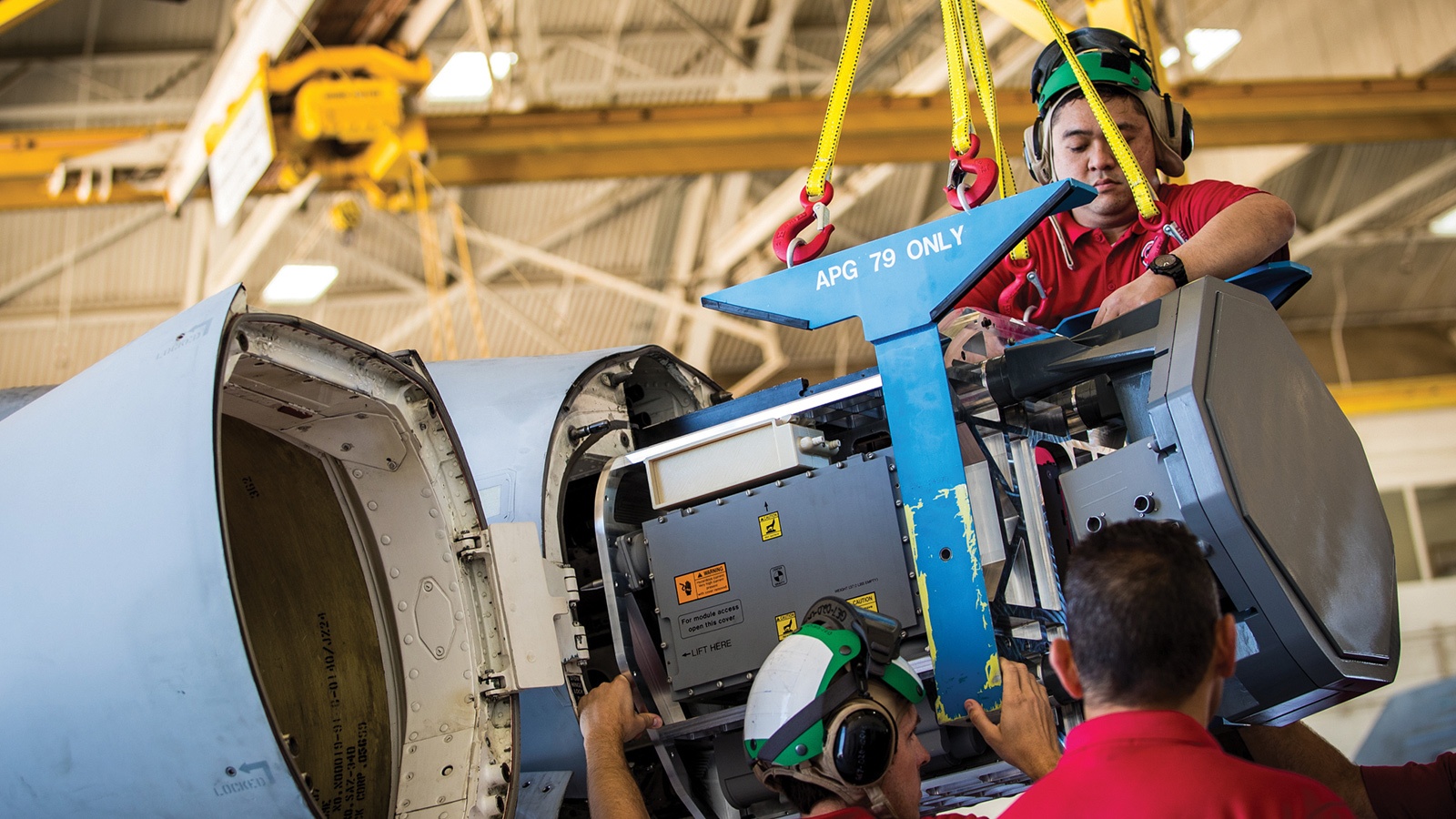 Technicians are installing equipment into the nose of an aircraft in a hangar, using a hoist labeled 