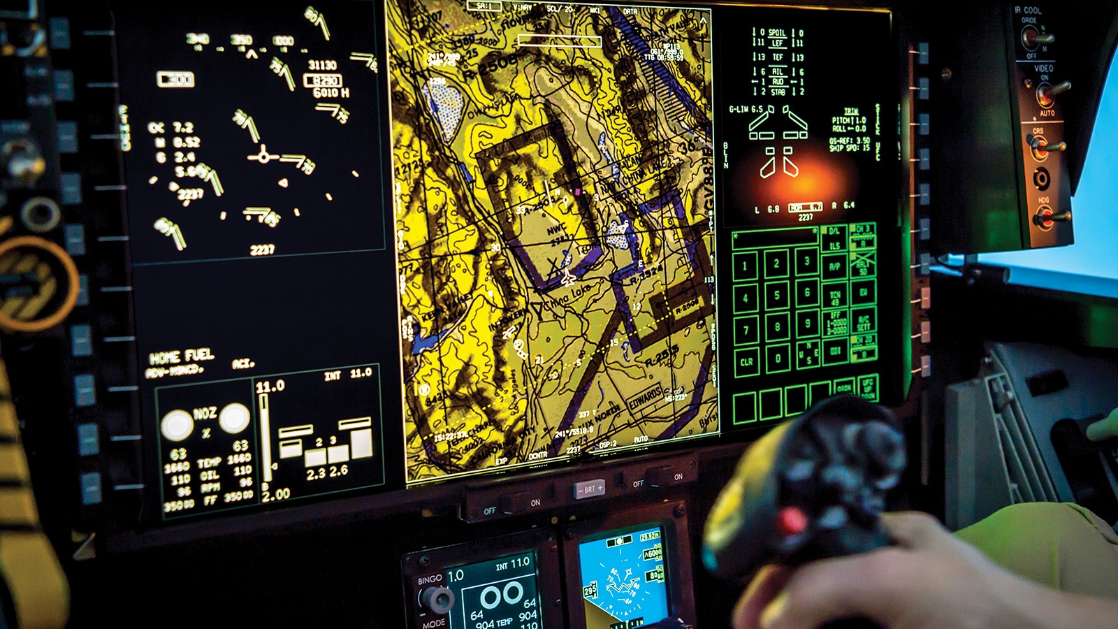 A pilot operates a joystick in front of a cockpit display showing a detailed topographical map and various flight data indicators.