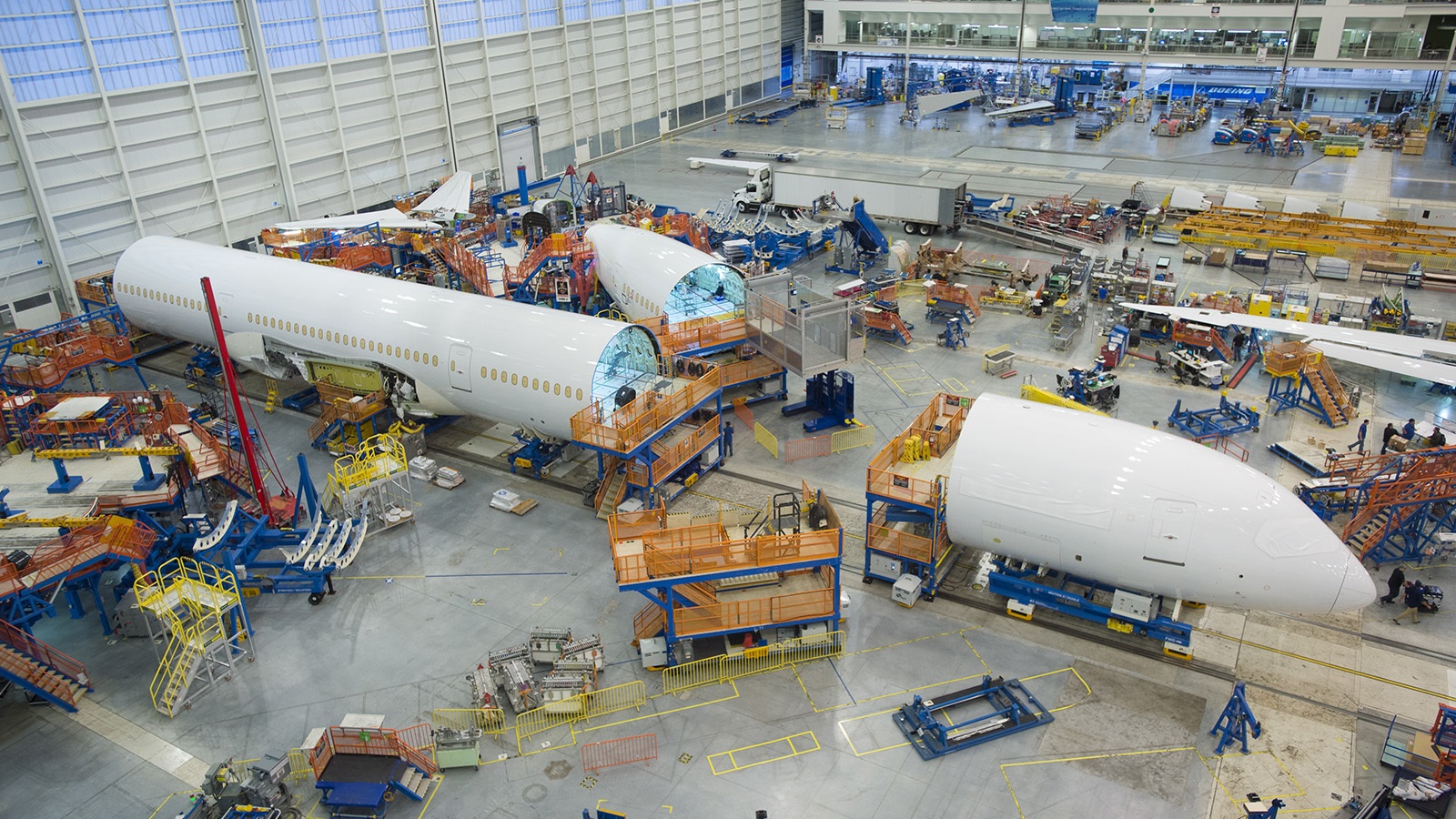 Inside a large aircraft assembly facility, a partially assembled airplane fuselage is surrounded by construction equipment and workers in a bustling, organized setup.
