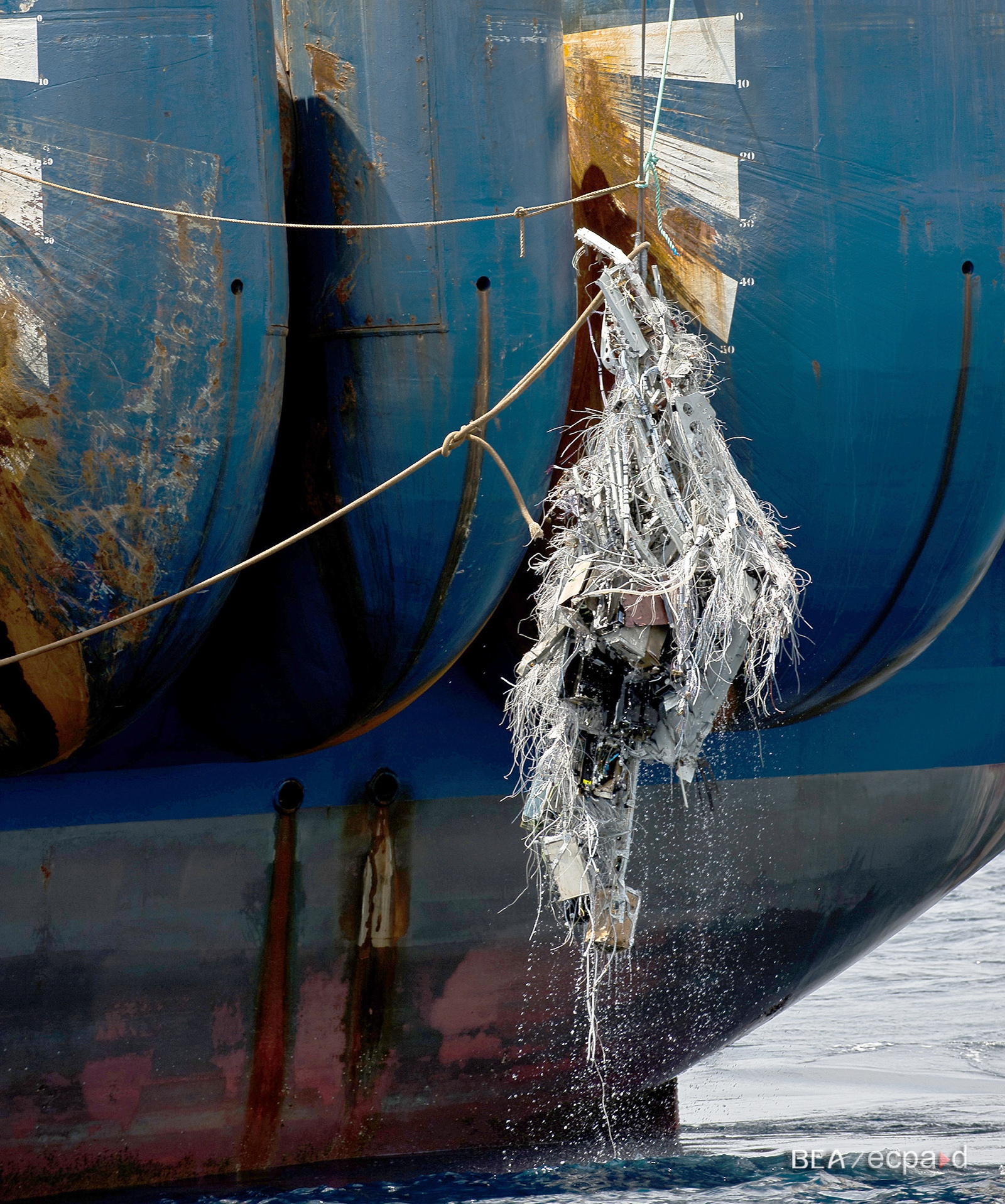 A large ship's hull partially submerged in water, with a crane hoisting a tangled mass of metal debris and cables dripping water.