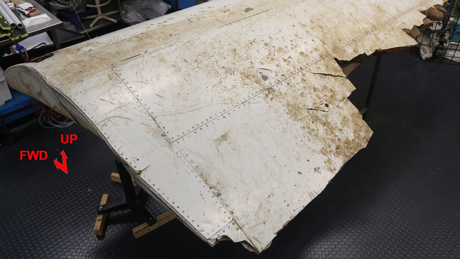 An aged, damaged aircraft wing section displayed on a stand in a workshop. Text labels indicate "FWD" and "UP" directions.