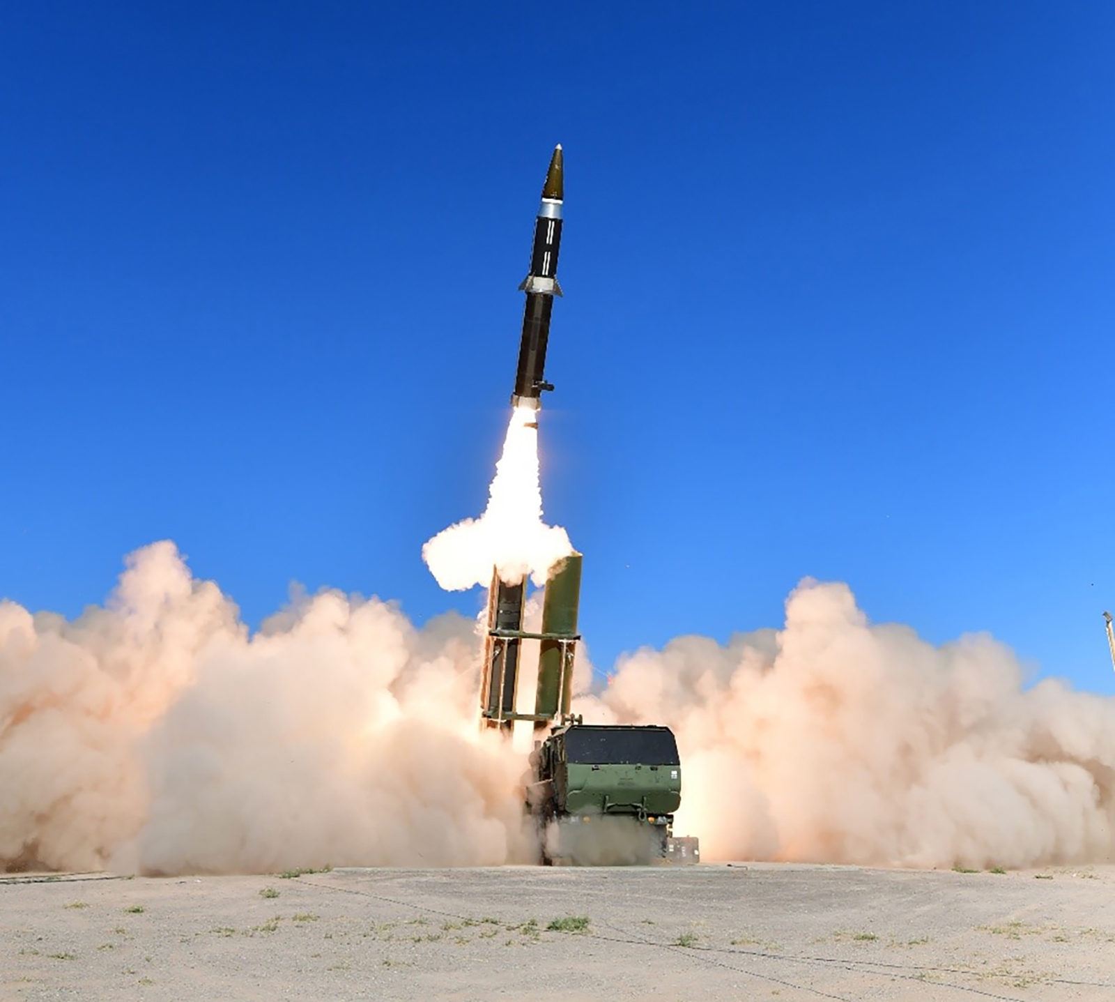A missile is being launched from a ground-based launcher against a clear blue sky, surrounded by clouds of dust and smoke.